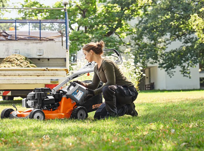 Eine Frau kniet auf dem Gras neben einem orangefarbenen Stihl Moto4plus Rasenmäher